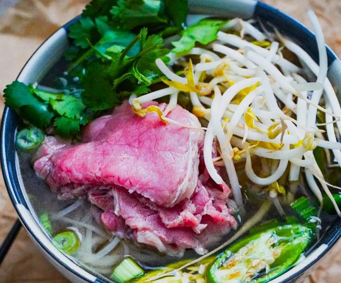 A vibrant bowl of pho featuring bright pink slices of steak, fresh sprouts, greens, and pickles.