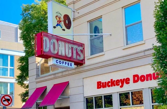 The storefront features a prominent sign for donuts, another for coffee, and displays the shop's name on the facade, along with an illustrated sign of a whimsical animal character in a chef's hat pushing a massive donut.