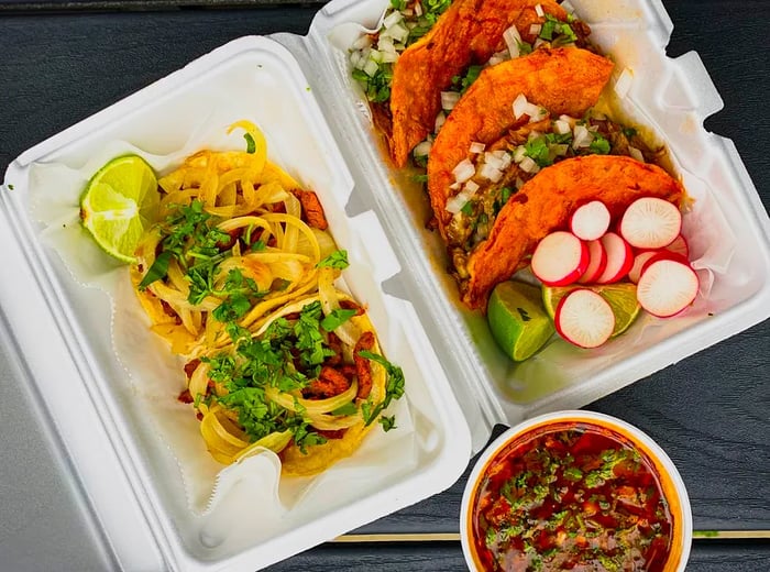 A stack of tacos in a plastic takeout container, garnished with sliced radishes and lime wedges, next to a paper cup filled with deep red broth.