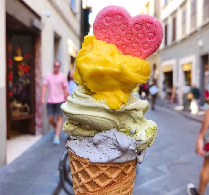 A whimsical gelato cone stacked high, adorned with an edible heart, set against a bustling street filled with people.