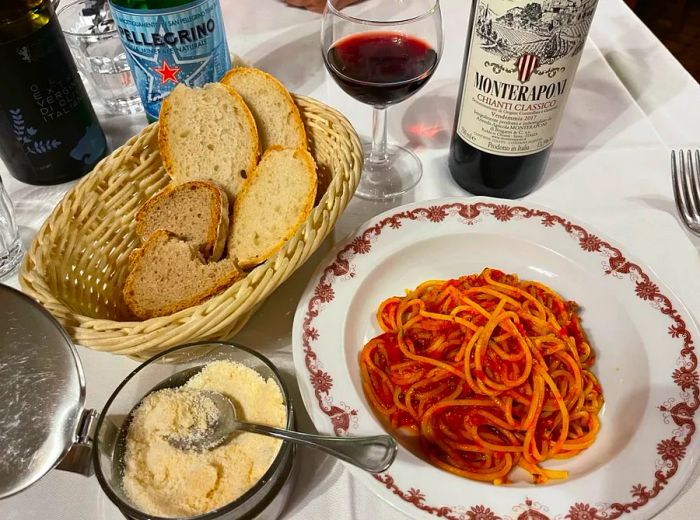 A table set with a plate of pasta, a basket of bread, grated cheese, and a bottle of red wine