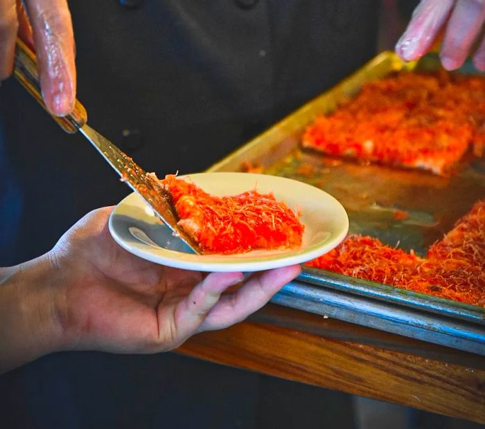 A gloved server carefully places a slice of knafi from a tray onto a plate using a metal spatula.