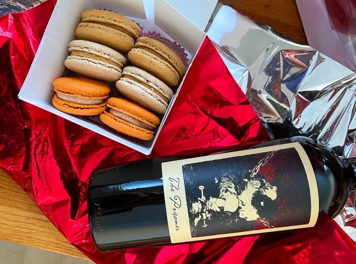 Aerial view of a paper box filled with macarons in an array of colors, next to a large bottle of red wine.