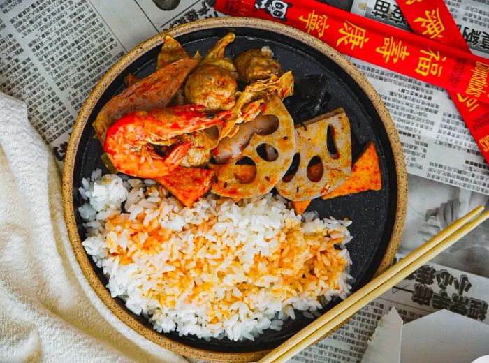 An overhead view of a shallow ceramic dish filled with fluffy white rice topped with vibrant red sauce, accompanied by a heap of fresh head-on shrimp, pickled vegetables, and small meatballs.