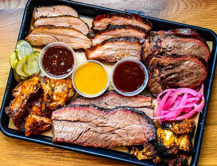 Aerial view of a large platter showcasing an array of barbecued meat slices, neatly arranged by type, accompanied by various dipping sauces.