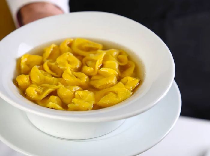 A server presents a bowl of tortellini in broth on a saucer.