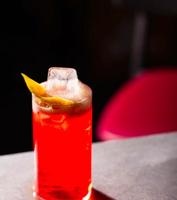 A vibrant red highball featuring a large ice cube and a lemon twist elegantly perched on top.