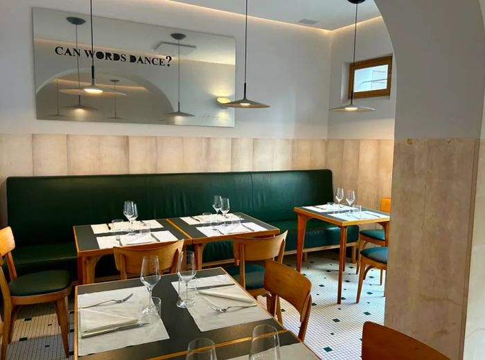 The restaurant features an interior adorned with sage green banquettes, wooden mid-century chairs, and classic tiled floors.