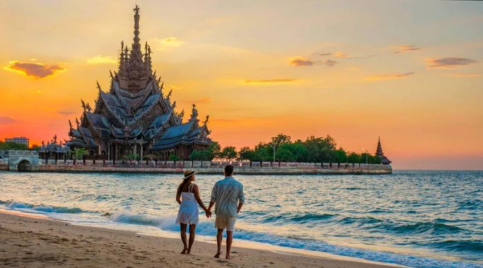 A couple strolls along the beach at Pattaya, with the Sanctuary of Truth temple in view.