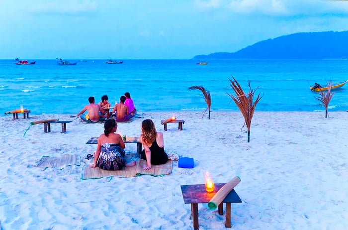 Tourists relax on a beach at sunset, with soft white sand and turquoise waters. Lanterns glow softly as twilight descends.