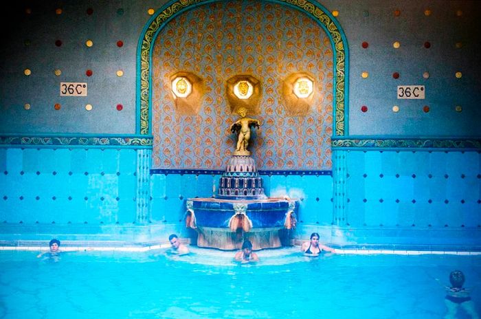 People relax in a steaming blue bath adorned with blue tiles next to an art nouveau fountain. A sign on the tiled wall indicates the water temperature is 36°C.