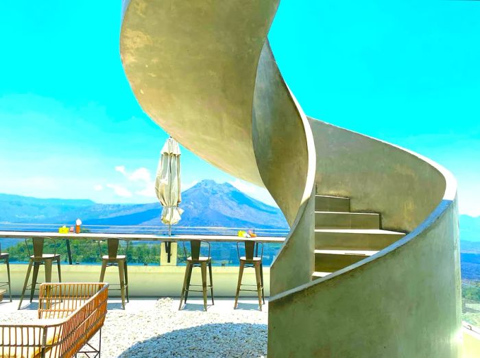 A concrete spiral staircase ascends to a café's outdoor terrace, offering stunning views of Mount Batur.