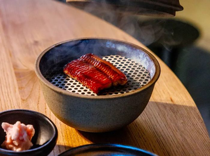 A lid is lifted from a mini grill, revealing glazed eel alongside pickled vegetables and another dish in the background.