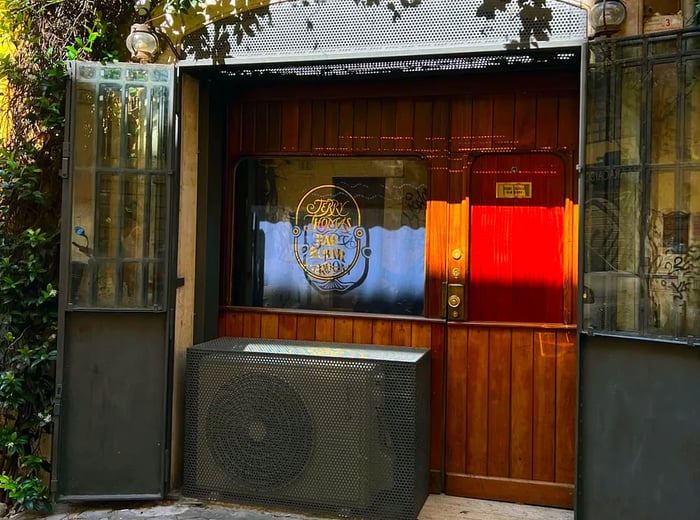 A wooden door nestled in a sunny alcove surrounded by greenery, next to a window featuring the bar's name in elegant cursive.