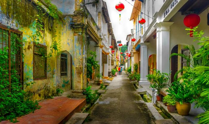 A narrow, historic street adorned with red Chinese-style paper lanterns strung between the buildings, as two figures walk away from the camera.