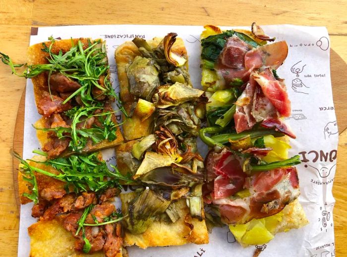 Square-cut pieces of pizza with an array of toppings arranged on wax paper on a tray.