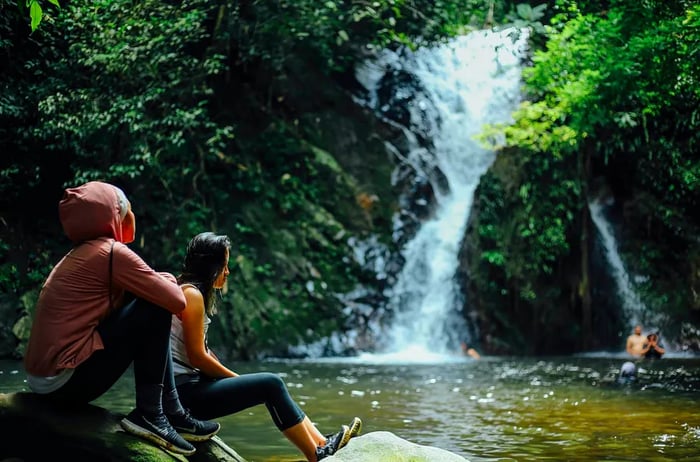 Two women relax by a pool beside a waterfall in the jungle