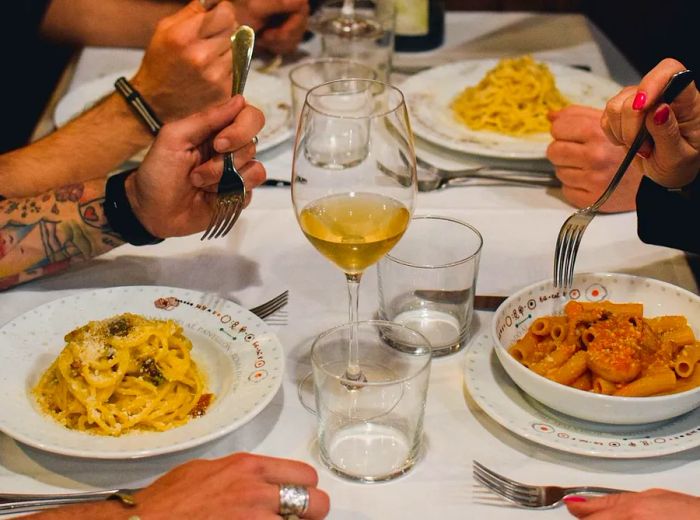 Four diners enjoy pasta and wine at a table adorned with a white tablecloth.