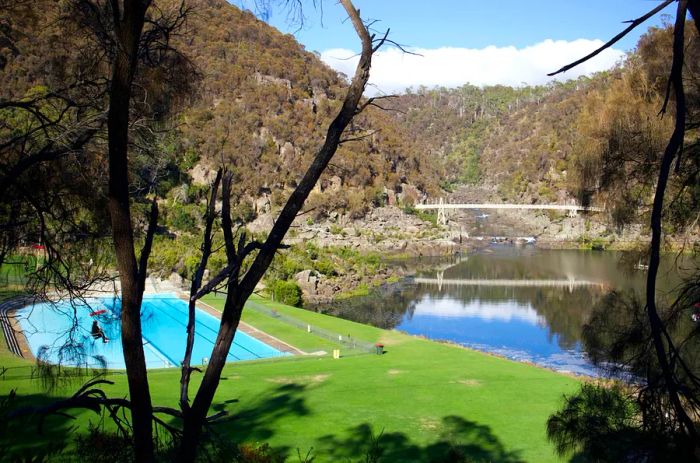 A picturesque scene showcasing a grassy area with a swimming pool, framed by trees and a peaceful river. In the background, a bridge and rolling hills enhance the tranquil landscape.