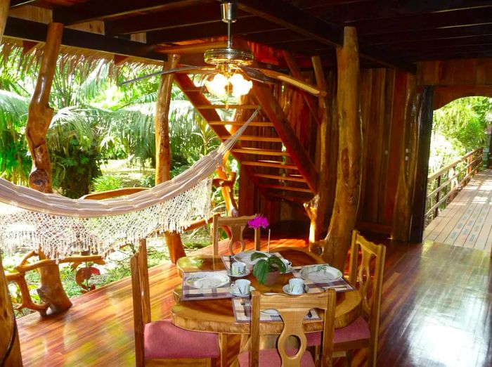 Interior view of the Topos Tree House showcasing its wooden floors and paneling, featuring a wooden table and chairs in the center with a white canopy to the left.