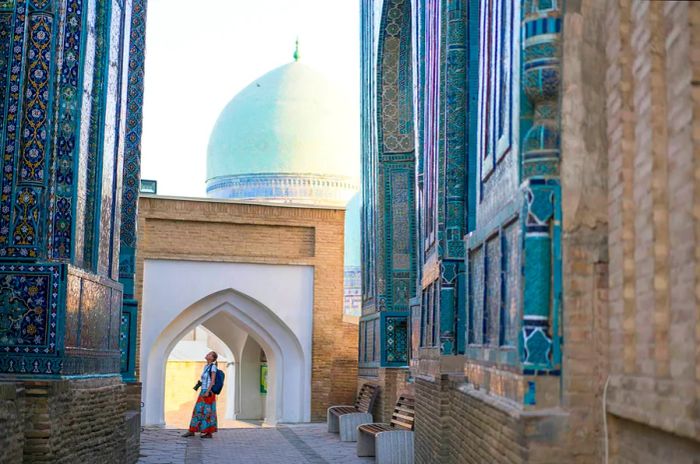 An elderly woman walks along the path connecting the memorial structures of the Shakhi Zinda Mausoleum, a complex showcasing Islamic architecture from the 9th to 12th centuries.