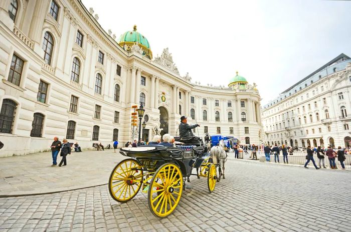 A fiaker, a horse-drawn carriage with a coachman serving as a tourist guide, passing by the Hofburg palace at Michaelerplatz, filled with tourists and locals.