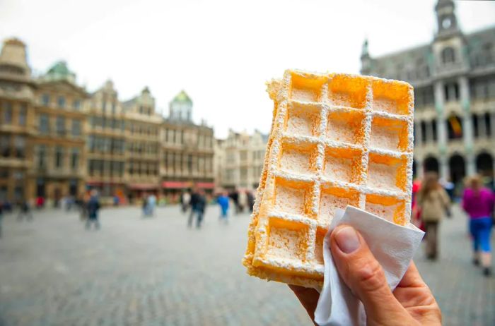 A hand holding a Belgian waffle sprinkled with powdered sugar in the Grand Place, Brussels