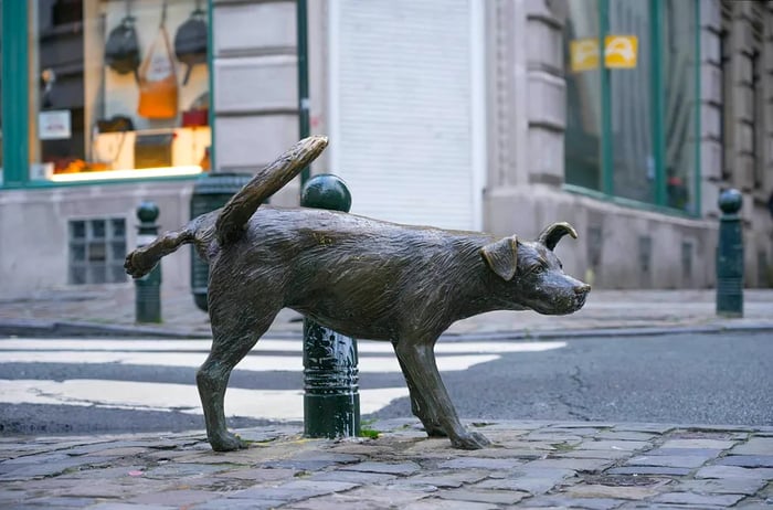 A bronze statue of a male dog lifting its leg beside a bollard on a street in Brussels, Belgium