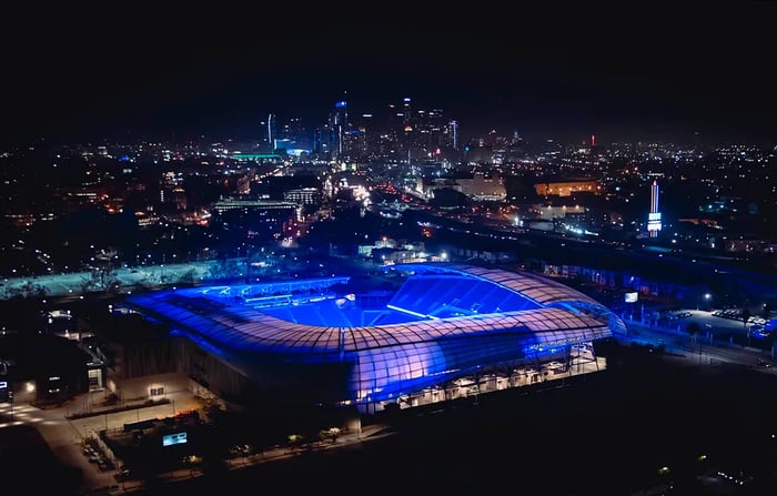 An aerial view of BMO Stadium glowing with purple lights, framed by the Los Angeles skyline in the background.