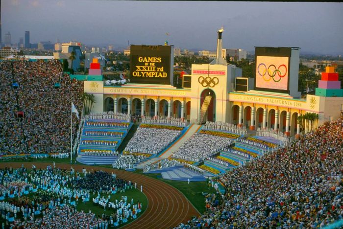 Rafer Johnson ignites the Olympic flame during the opening ceremony of the Los Angeles games, captured at the top of the steps, dressed in white, right beneath the Olympic rings.