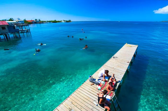 Girls play music on a sunlit dock in Utila, with snorkelers enjoying the blue-green waters behind them.