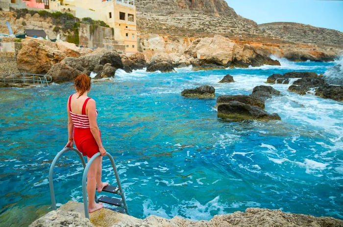 A woman in a red swimsuit stands by the ladder leading into an inlet at Għar Lapsi, surrounded by rocky cliffs in Malta.