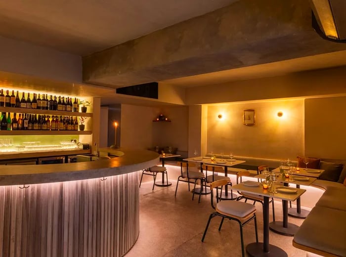 An elegant bar interior featuring tables along a plush banquette, softly lit ambiance, a prominent backlit bar, and an abundance of pink and gold accents.