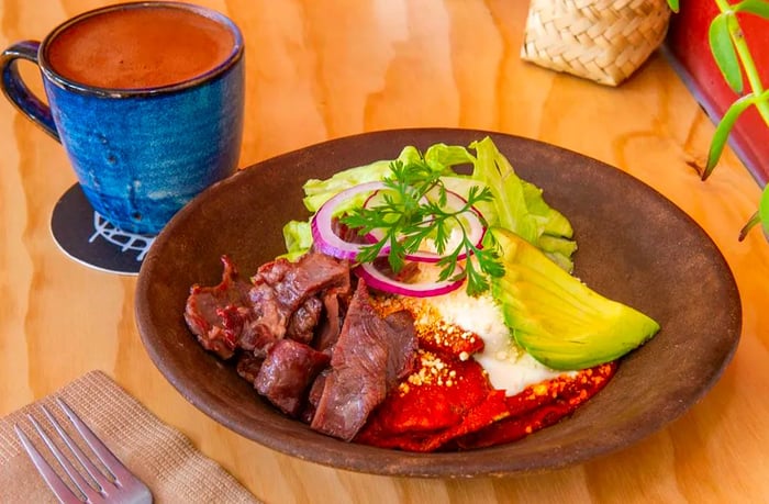 A plate featuring sliced meat, avocado, onions, fresh herbs, and salsa, paired with a cup of coffee.