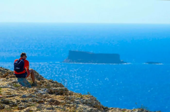 A male hiker takes a break on the rocky trail along the Dingli Cliffs, gazing out at Filfla Island, Malta.