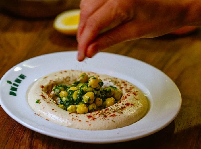 A chef adds spices to a plate of hummus garnished with whole chickpeas.