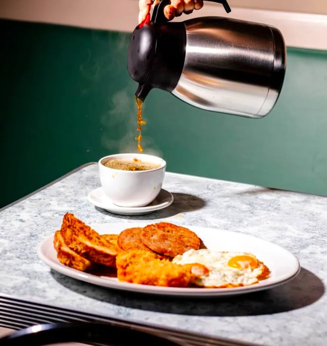 A hand, unseen, fills a mug with coffee next to a plate of assorted breakfast foods.