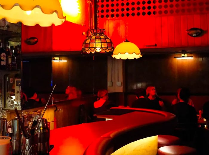 A dimly lit bar with dark red hues, featuring antique pendant lights above a curved wooden bar and diners seated in shadowy booths.