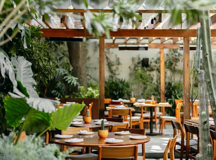 A restaurant patio surrounded by greenery, featuring blond wood tables, chairs, and a cozy canopy.