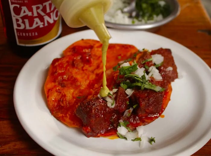 A hand drizzles green sauce from a squeeze bottle onto a juicy, saucy taco.