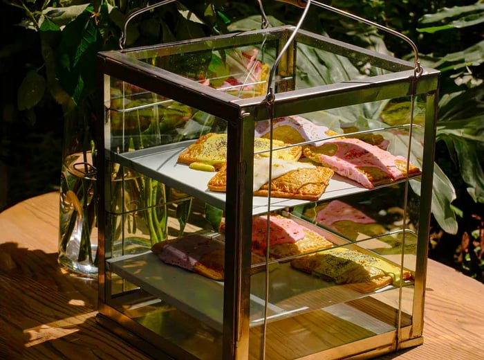 A glass pastry display case with handles, set against a backdrop of greenery.