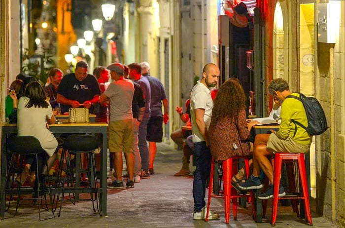 People enjoy drinks at tables outside a bar nestled in a narrow street in Valletta, Malta.