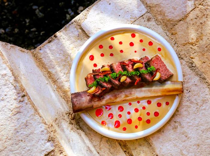 An aerial view of a plate resting on stone steps, showcasing a large short rib with the bone, drenched in a creamy sauce and dotted with chile oil.