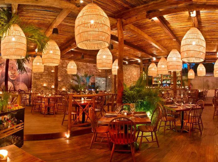 A spacious, unoccupied dining room adorned with natural wood, featuring a stone wall at the back, large pop-art decorations, woven lanterns hanging from above, and tables elegantly set for dinner.