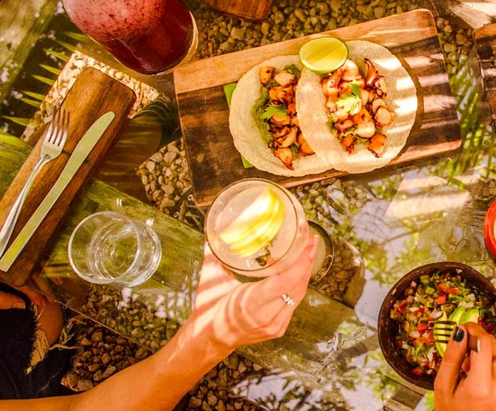 From an aerial view, guests are seen relishing a variety of snacks and beverages arranged on a glass table set atop pebble flooring.