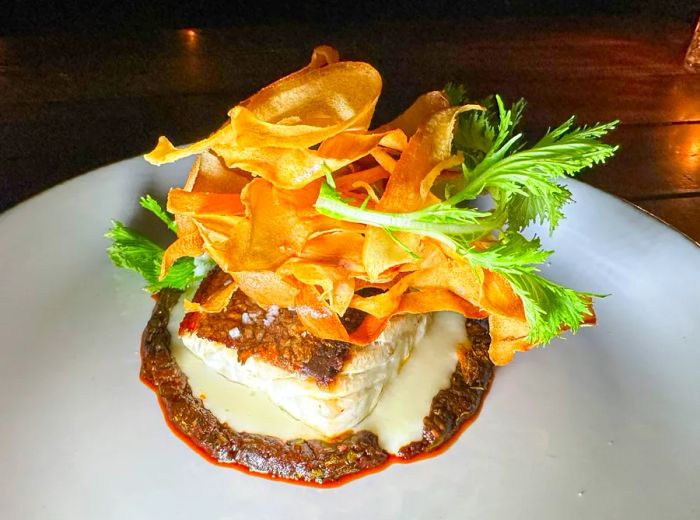 A plate showcasing a piece of white fish crowned with an elaborate tower of crunchy toppings and greens.