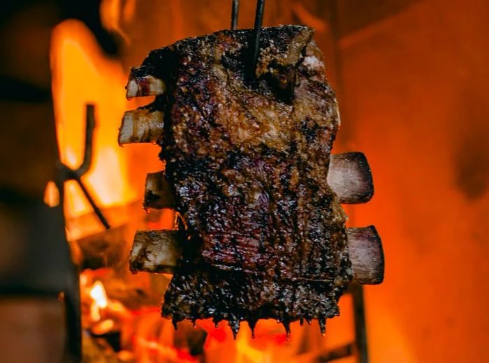 A rack of ribs dangles from a metal hook above a roaring fire.