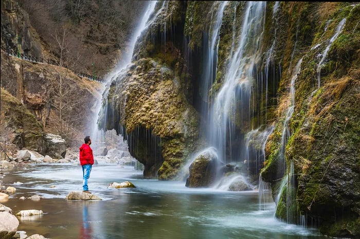 A man dressed in red rain gear and hiking boots perches on a rock above a pool, gazing at a waterfall.