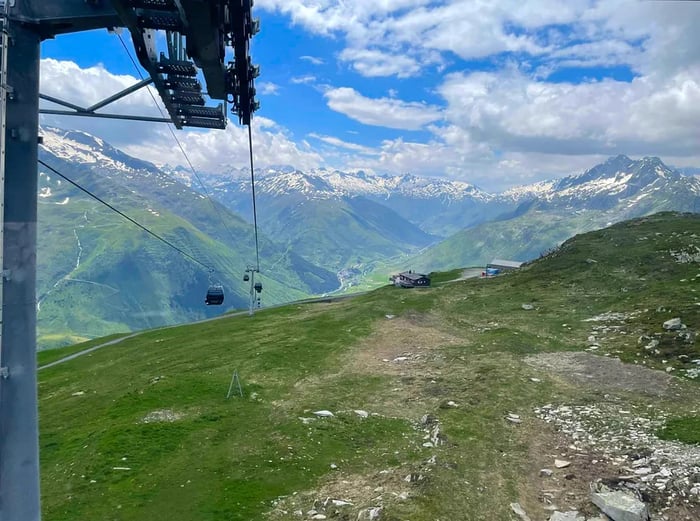 A gondola ascends the mountain, surrounded by lush green hills and towering snow-capped peaks.