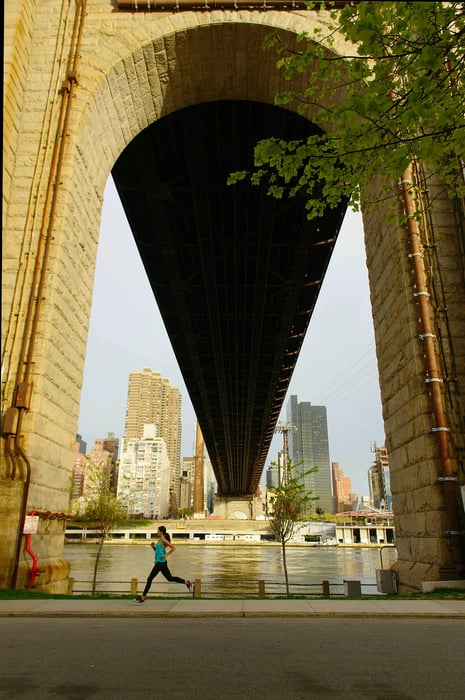 A woman jogs along the sidewalk beneath one of the arches of the Queensboro Bridge on Roosevelt Island, New York, with skyscrapers and large buildings visible across the river.